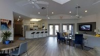 This single-floor community space offers an open-concept layout designed for comfort and social interaction. Upon entry, a prominent "Welcome" sign sets a friendly tone, leading into a spacious main area with vaulted ceilings and multiple ceiling fans that enhance airflow and comfort throughout. The kitchen area is fully equipped with stainless steel appliances, including a refrigerator and microwave, and features a long bar counter lined with sleek black bar stools — ideal for casual gatherings or quick refreshments. The lounge area is thoughtfully furnished with navy blue upholstered chairs and a sofa accented with yellow throw pillows, centered around a glass-top coffee table and anchored by a patterned area rug. A stylish floor lamp adds a warm, ambient glow to the seating arrangement. Wide French doors line the far wall, flooding the interior with natural light and providing views of an outdoor patio area. The flooring throughout is a warm, medium-toned hardwood-style material that ties the space together cohesively. Recessed lighting supplements the natural light, ensuring the space remains bright and inviting at all hours. The overall ambiance is modern, clean, and welcoming — well-suited for community events and everyday use.