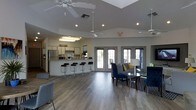 This single-floor community space offers an open-concept layout designed for comfort and social interaction. Upon entry, a prominent "Welcome" sign sets a friendly tone, leading into a spacious main area with vaulted ceilings and multiple ceiling fans that enhance airflow and comfort throughout. The kitchen area is fully equipped with stainless steel appliances, including a refrigerator and microwave, and features a long bar counter lined with sleek black bar stools — ideal for casual gatherings or quick refreshments. The lounge area is thoughtfully furnished with navy blue upholstered chairs and a sofa accented with yellow throw pillows, centered around a glass-top coffee table and anchored by a patterned area rug. A stylish floor lamp adds a warm, ambient glow to the seating arrangement. Wide French doors line the far wall, flooding the interior with natural light and providing views of an outdoor patio area. The flooring throughout is a warm, medium-toned hardwood-style material that ties the space together cohesively. Recessed lighting supplements the natural light, ensuring the space remains bright and inviting at all hours. The overall ambiance is modern, clean, and welcoming — well-suited for community events and everyday use.