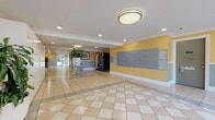 Located at 1600 NE 135th St in North Miami, Florida, this building lobby offers a welcoming and well-maintained common area designed for everyday resident use. The expansive entrance is finished with large-format cream and beige diagonal-set ceramic tile flooring, creating a clean and polished aesthetic throughout. Warm yellow accent walls trimmed with white crown molding and a two-tone gray wainscoting give the space a bright, cheerful character, while recessed lighting and flush-mount ceiling fixtures ensure consistent illumination.

Floor-to-ceiling glass windows and automatic sliding entry doors flood the space with abundant natural light and provide clear views of the surrounding landscaping and parking area. Lush potted tropical plants, including large leafy arrangements displayed on a decorative pedestal, add a fresh, inviting touch to the interior.

A prominent bank of resident mailboxes lines one wall, providing convenient centralized mail access. A vending machine is positioned nearby for added convenience. The corridor connects the main entry to elevator banks finished with tiled surrounds, facilitating easy movement throughout the building. A seating bench near the entry offers a comfortable waiting area. The overall ambiance is clean, functional, and welcoming, reflecting a well-kept residential building environment.