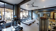 This fitness center is thoughtfully designed to provide a motivating and functional workout environment. The space features a curated selection of modern cardio equipment, including a row of treadmills and elliptical trainers from True Fitness, catering to a range of exercise preferences. Architectural character is added through dark exposed beam accents that contrast beautifully against the crisp white ceiling, while the bold blue-gray walls create an energizing yet focused ambiance. Large floor-to-ceiling windows line one wall, flooding the space with natural light and offering views to the outdoors, enhancing the open and airy feel of the room. Ceiling fans are strategically positioned overhead to maintain comfortable airflow throughout workouts. A mirrored section along one wall visually expands the space and allows users to monitor their form during exercise. The flooring is finished in a durable, cushioned material appropriate for fitness use. The overall design balances industrial-inspired aesthetics with practical amenities, resulting in a polished, well-appointed fitness facility that is both inviting and fully functional for daily use.