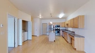 This single-floor space offers a bright and functional open-concept layout with light hardwood flooring that extends seamlessly throughout the main living area. The kitchen serves as the focal point of the space, designed with warm light wood cabinetry, dark granite countertops, and a full suite of stainless steel appliances, including a refrigerator, microwave, and range. A central kitchen island provides additional prep space and doubles as a casual gathering spot, illuminated by pendant lighting above. The countertops continue along an L-shaped configuration, incorporating a double sink beneath additional upper cabinetry that maximizes storage. The open layout allows for a generous living area that flows naturally from the kitchen, offering flexibility in how the space can be arranged and utilized. A dedicated in-unit laundry alcove is conveniently tucked to one side, housing a full-size washer and dryer. The space benefits from high ceilings that contribute to an airy, expansive feel. Neutral wall tones and consistent flooring throughout create a cohesive and clean aesthetic. Multiple doorways provide access to additional rooms, completing this well-organized and practical floor plan.