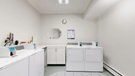 This shared laundry facility is designed with functionality and cleanliness in mind, finished throughout in crisp white tones with light green-tinted tile flooring. The main laundry room houses multiple front-loading and top-loading commercial washers and dryers, each equipped with card or coin payment interfaces, making laundry tasks straightforward and efficient. A built-in white laminate folding counter with cabinet storage runs along one wall, providing ample space for sorting and folding laundry. An octagonal mirror is mounted above the counter, adding a subtle decorative touch to the otherwise utilitarian space. A wall clock and posted usage instructions ensure users stay informed. Overhead fluorescent lighting keeps the room bright, while a baseboard heater maintains comfortable temperatures. The facility is accessed via a well-maintained corridor finished with the same tile flooring and neutral walls. The hallway features a fire hose cabinet, fire alarm pull station, and clearly marked emergency exit signage, reflecting strong safety standards. An elevator nearby provides convenient, accessible entry to the facility. The overall environment is orderly, hygienic, and practical, catering effectively to the needs of building residents.