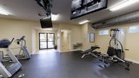 This fitness center offers a practical and well-organized space designed to support a full range of exercise activities. The main workout area features durable rubber flooring throughout, providing a safe and functional surface for all types of training. The warm, neutral-toned walls and bright fluorescent lighting create a clean, focused environment conducive to exercise.

The cardio zone is equipped with multiple treadmills, elliptical trainers, and a stationary bike, giving users a variety of aerobic options. Multiple flat-screen televisions are ceiling-mounted throughout the space, allowing members to stay entertained during workouts. A dedicated strength training area includes a multi-function cable machine, an adjustable weight bench, a set of free weights, and additional resistance equipment.

A built-in dual water fountain station provides convenient hydration access directly within the facility. Exposed HVAC ductwork along the upper walls ensures proper ventilation throughout the space. Natural light filters in through a high horizontal window, complementing the overhead lighting. Glass-panel entry doors open to the exterior, connecting the facility to the surrounding outdoor area. An interior hallway leads to additional ancillary spaces. A wall-mounted clock and informational postings round out the functional amenities of this comprehensive fitness facility.