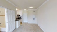 This single-floor unit presents an open-concept design that visually connects the living area and kitchen, creating a cohesive and functional layout. The living space is finished with plush, neutral-toned carpeting that adds warmth underfoot, while the kitchen and entryway transition to hard flooring for practicality. Crown molding runs along the ceiling throughout, adding a refined architectural touch to the otherwise clean, minimal aesthetic.

The kitchen is positioned along one wall and features white cabinetry, dark countertops, and stainless steel appliances including a range and an over-the-range microwave. A breakfast bar-style counter separates the kitchen from the living area, providing additional workspace and a casual dining option.

Flush-mount ceiling light fixtures illuminate both the living space and the entryway, ensuring consistent, even lighting throughout. The front entry door is white and centrally positioned, flanked by a utility panel and an HVAC access panel built into the wall. The overall palette—white trim, cream walls, and light carpet—creates a bright, neutral environment that is move-in ready and easy to personalize.