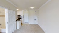 This single-floor unit presents an open-concept design that visually connects the living area and kitchen, creating a cohesive and functional layout. The living space is finished with plush, neutral-toned carpeting that adds warmth underfoot, while the kitchen and entryway transition to hard flooring for practicality. Crown molding runs along the ceiling throughout, adding a refined architectural touch to the otherwise clean, minimal aesthetic.

The kitchen is positioned along one wall and features white cabinetry, dark countertops, and stainless steel appliances including a range and an over-the-range microwave. A breakfast bar-style counter separates the kitchen from the living area, providing additional workspace and a casual dining option.

Flush-mount ceiling light fixtures illuminate both the living space and the entryway, ensuring consistent, even lighting throughout. The front entry door is white and centrally positioned, flanked by a utility panel and an HVAC access panel built into the wall. The overall palette—white trim, cream walls, and light carpet—creates a bright, neutral environment that is move-in ready and easy to personalize.