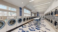Located at 1 Marion Pepe Dr in Lodi, NJ, this modern laundromat offers a clean, well-organized single-floor facility designed for efficiency and comfort. The interior features an expansive main washing area lined with rows of front-load commercial washing machines, including large-capacity Speed Queen units, arranged along both walls to maximize usable floor space. A dedicated dryer section with stacked commercial dryers runs along the opposite wall, providing ample drying capacity. The space is visually distinguished by a bold blue and white chevron-patterned tile floor, complemented by navy blue accent panels on the walls that add a contemporary aesthetic. Vaulted ceilings with recessed lighting and long linear LED fixtures ensure the space is brightly and evenly lit throughout. A series of clerestory windows along the upper walls invite natural light and views of the surrounding neighborhood. The facility includes centrally placed folding tables with chrome laundry carts for customer convenience. A comfortable waiting area near the entrance is furnished with sleek navy blue chairs, a glass coffee table, and a wall-mounted flat-screen television. The front entrance features glass double doors that open to the exterior, further enhancing the bright and welcoming atmosphere of this well-appointed laundry facility.