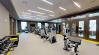 This contemporary fitness center is designed to support a comprehensive workout experience across multiple dedicated zones. The space is anchored by a spacious main floor finished with neutral-toned carpet and bold yellow accent flooring trim, creating a clean, energetic aesthetic throughout.

The cardio zone lines one wall, outfitted with a row of treadmills and elliptical machines, each equipped with individual screens. Large windows along this section fill the area with natural light, while a wall-mounted flat-screen TV adds an entertainment option for users. Full-length mirrors amplify the sense of space and allow exercisers to monitor their form.

An extensive strength training area occupies the central and adjacent sections of the floor, housing a broad selection of commercial-grade cable and resistance machines. A separate room, accessible through glass-paneled doors, provides additional equipment including a dumbbell rack and supplementary strength machines.

A standout feature of this fitness center is its dedicated boxing zone. A textured brick accent wall branded with "Brandon X-066 Boxing Club Ringside" sets the tone, and the area is equipped with punching bags, speed bags, and boxing accessories, giving the space a distinct, athletic character.

Recessed linear LED lighting runs across the ceilings throughout, providing consistent, bright illumination. Restroom facilities are conveniently located on-site, rounding out this fully self-contained fitness facility.