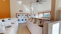 This communal laundry facility is thoughtfully designed to accommodate a high volume of use while maintaining a clean, comfortable environment. The room features soaring vaulted ceilings that create an open, airy atmosphere, complemented by a large ceiling fan that promotes air circulation throughout the space. Warm natural light streams in through wood-framed windows, offering pleasant outdoor views and reducing the need for artificial lighting during the day. Recessed lighting is also present to ensure the space remains well-lit at all times.

The facility is outfitted with a generous row of top-loading washing machines aligned along one side, providing ample capacity for simultaneous loads. Stacked dryer units are arranged along the back wall, maximizing the use of vertical space efficiently. The machines are uniformly white and well-maintained, contributing to the room's clean and orderly appearance. The flooring features a speckled terrazzo-style surface that is both durable and easy to keep clean. HVAC vents are positioned throughout the upper walls to maintain a comfortable temperature year-round. Overall, the space combines practical functionality with a bright, welcoming design.