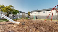 Located at 809 Dozer Dr in West Jefferson, Ohio, this outdoor playground area serves as a communal recreational space within a residential community. The ground-level space is surfaced with wood chip mulch, providing a soft, safety-conscious base throughout the play area. Key features include a red A-frame swing set, a freestanding slide with metal stair access, and a jungle gym climbing structure, offering a variety of play options for children of different ages and abilities. Multiple park-style benches are positioned around the perimeter, providing comfortable seating for supervising adults. The area is enclosed by a wooden privacy fence, creating a defined and secure boundary. Surrounding residential buildings with light-colored siding and pitched, dark shingled roofs frame the space on multiple sides, reinforcing its neighborhood setting. Patches of grass and mature trees are visible along the edges of the play area, adding natural greenery to the environment. A lamppost is also present, suggesting the area may be accessible during early morning or evening hours. The open layout allows for clear sightlines across the entire play space, and the setting benefits from ample open sky, providing plenty of natural light throughout the day.