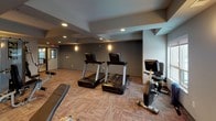 This fitness center offers a comprehensive workout environment thoughtfully designed to accommodate a range of exercise needs. The space features a tray ceiling with recessed lighting and wall-mounted sconces that create a well-lit, comfortable atmosphere. Neutral gray walls and patterned carpet flooring contribute to the modern, clean aesthetic throughout.

The cardio area is equipped with multiple Cybex treadmills, an elliptical trainer, and a stationary bike, positioned near large windows that allow natural light to filter in and provide views of the outdoors. A dedicated strength training zone houses a multi-station cable machine along with various resistance-based equipment, offering options for full-body workouts.

A free weight section lines one wall, featuring a well-stocked dumbbell rack alongside wall-mounted mirrors and flat-screen televisions, creating a functional and engaging workout environment. A water fountain is conveniently located near the exit, adding to the practical amenities of the space. The layout is open and organized, allowing for comfortable movement between equipment zones, and the space is accessible via a glass-paneled door.