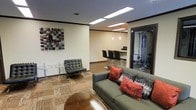 This single-floor space presents a thoughtfully designed interior that blends comfort with functionality. The main lounge area features a contemporary gray sofa accented with rich red and patterned throw pillows, complemented by a sleek leather Barcelona-style chair and a round side table — creating an inviting seating arrangement suited for relaxation or socializing. A mixed-material area rug anchors the lounge, adding texture and warmth to the hardwood-style flooring beneath.

Overhead, recessed rectangular light fixtures provide consistent, even illumination throughout the space. The walls are neutral in tone, with a bold, colorful mosaic-style wall art piece serving as a focal point in the lounge area. Crown molding adds a refined architectural detail along the ceiling edges.

Transitioning toward the back, a bar-height counter with black stools and pendant lighting above creates a casual dining or work nook, alongside what appears to be a small countertop appliance station. A glass-paneled door on the right side opens to a fitness area, hinting at gym equipment within. The overall ambiance is modern, clean, and multi-functional.