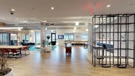 The lobby at 1 Alexander Street North in Yonkers, New York is a thoughtfully designed, single-floor space that blends industrial and contemporary aesthetics to create a welcoming and functional environment. Entry is made through sleek, black-framed glass doors that open into a bright, open interior finished with wood-look tile flooring throughout.

The main gathering area is expansive and airy, anchored by exposed concrete columns, suspended globe pendant lighting, and visible ductwork along the ceiling that lend an industrial character to the space. A full-length shuffleboard table serves as a central social feature, complemented by lounge seating with vibrant potted plants and a wall-mounted television. Along the perimeter, communal bench seating and bar-height tables provide additional gathering options beside large windows that invite natural light and outdoor views.

A staffed reception desk finished in dark metal with a warm wood counter top anchors one section of the lobby, accompanied by a digital display panel and pendant lighting overhead. Nearby, an open metal shelving unit divides areas while maintaining visual connectivity across the floor.

More intimate zones include a cozy seating alcove with wingback chairs beneath a warm wood-paneled ceiling, a small private meeting nook with a round table and mid-century style chairs, and a quiet lounge corner with a teal accent rug and modern furnishings. Restrooms are also conveniently accessible within the lobby level.