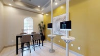 This single-floor business center offers a thoughtfully designed environment that balances functionality with contemporary style. The space is anchored by a bright, open lobby featuring large arched glass entry doors, light tile flooring throughout, and recessed lighting complemented by elegant chandelier fixtures. A central round accent table with decorative floral arrangement serves as a welcoming focal point upon entry.

The main common area provides multiple lounge seating arrangements, including plush armchairs and dark upholstered sofas, separated by structural columns that define distinct zones without sacrificing the open feel of the space. Curated artwork and indoor greenery add warmth and visual interest throughout.

A dedicated workstation area features sleek pedestal-mounted computer terminals with compact round surfaces, positioned against bold yellow accent walls that energize the space. Framed abstract artwork adds a refined touch to this productive zone.

Adjacent to the workstations, a high-top meeting table surrounded by upholstered bar-height chairs provides a collaborative setting for small group work or informal meetings. A graceful arched window floods this nook with natural light and offers views of the surrounding landscape, further enhancing the inviting atmosphere of this well-equipped business center.