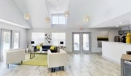 Located at 415 Prestwick Ridge Way in Knoxville, Tennessee, this single-story community clubhouse is designed to impress with its airy, contemporary aesthetic and welcoming atmosphere. The centerpiece of the space is a grand open lounge area distinguished by soaring vaulted ceilings that amplify the sense of volume and openness throughout. Large windows fitted with clean white blinds allow abundant natural light to flood the interior, complementing the neutral light gray walls and light-toned wood-look flooring that runs throughout the space. A series of decorative globe pendant lights hang elegantly from the ceiling, serving as both functional lighting and striking design focal points. The lounge is furnished with a modern seating arrangement, including a deep navy blue sofa accented with bold yellow throw pillows, white accent chairs, and a gold-framed glass coffee table set atop a chartreuse area rug — creating a vibrant yet cohesive color palette. Flanking table lamps and decorative vases add refined finishing touches. A set of glass-paneled double doors provides direct access to an outdoor area, inviting natural connection to the exterior surroundings. The overall ambiance is bright, polished, and inviting, making this clubhouse a well-appointed gathering space for the community.
