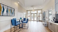 This single-floor space functions as a well-appointed lobby and communal gathering area, thoughtfully designed to balance welcoming aesthetics with practical functionality. The entrance is defined by tall, glass-paneled double doors flanked by transom windows that flood the interior with generous natural light, creating an open and airy atmosphere throughout.

The main lobby area features light hardwood-style flooring that extends seamlessly across the space, complementing the neutral warm-beige wall tones and crisp white wainscoting panels. Soaring ceiling heights amplify the sense of openness, with recessed lighting and a classic pendant light fixture providing layered illumination.

A casual seating area anchors one side of the lobby, furnished with a round bistro-height table and coordinating blue upholstered chairs with sleek metal frames. Adjacent to this, a built-in credenza finished in muted blue cabinetry houses beverage and media amenities, topped with a mounted display screen.

A dedicated office area with a dark wood desk and guest seating is accessible from the lobby, separated by a glass-panel sliding door. A secondary corridor leads to additional rooms, including what appears to be a kitchenette or break space. Glass French doors open directly to an outdoor pool area, connecting the interior to the exterior seamlessly.