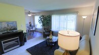 This single-floor residence showcases an open-concept layout that seamlessly connects the living and dining areas, creating a cohesive and functional flow throughout the main living space. The living area is anchored by a plush, dark area rug and features a glass-topped coffee table, a patterned accent chair, and a floor lamp that contributes warm ambient lighting. A large window fitted with vertical blinds allows natural light to filter into the room while offering a view of the greenery outside, enhancing the bright and airy feel of the space. A tall indoor plant near the window adds a refreshing natural element to the interior. Adjacent to the living area, the dining space accommodates a classic dining set with wrought-iron style chairs, suitable for everyday meals or casual entertaining. The dining area benefits from its own ceiling fan with an integrated light fixture, providing both ventilation and illumination. The flooring transitions from carpet in the living area to a harder surface in the dining zone, subtly defining each space. Overall, the interior presents a comfortable and practical environment with a relaxed, lived-in ambiance.