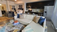 This single-floor space showcases a thoughtfully designed open-concept layout that seamlessly integrates the living, sleeping, and kitchen areas into one cohesive environment. The living area is anchored by a large white sectional sofa adorned with navy blue and gold geometric-patterned throw pillows, complemented by a glass-top coffee table and a patterned area rug that adds visual texture to the floor. The sleeping area is visible directly behind the living space, featuring a well-appointed bed with coordinating navy and gold bedding, maintaining a consistent color palette throughout. The kitchen occupies a dedicated nook along the back wall, outfitted with dark espresso cabinetry, stainless steel appliances including a microwave, stove, and dishwasher, and a neutral tile backsplash. A compact breakfast bar with counter seating bridges the kitchen and living areas, providing a functional dining and prep surface. The overall ambiance is contemporary and curated, with a rich navy, gold, and white color scheme lending a polished, cohesive aesthetic throughout the space.