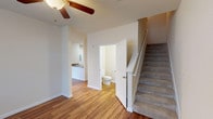 This two-story residence in Spring, Texas offers a well-organized layout across two floors, providing clear separation between living and sleeping areas. The main level features warm wood-look flooring throughout the open living and dining areas, with a ceiling fan overhead contributing to comfortable air circulation. A conveniently located half-bathroom on the first floor serves guests, while the galley-style kitchen is equipped with light wood cabinetry, dark granite-style countertops, black appliances including a refrigerator and electric range, and a stainless steel sink. A carpeted staircase leads to the upper level, where two bedrooms are situated, each featuring plush carpet flooring and natural light from windows with blinds. The bedrooms are served by two full bathrooms, both appointed with warm wood-toned vanities, light countertops, tile flooring, and walk-in shower or tub configurations. Additional storage is provided through a walk-in closet with wire shelving. The neutral warm-toned walls throughout create a cohesive, move-in-ready ambiance well-suited to a variety of décor styles.