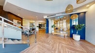 This thoughtfully designed lobby makes an immediate impression with its expansive, light-filled interior anchored by a dramatic arched glass entryway that floods the space with natural light. The herringbone-patterned wood flooring runs throughout, lending a refined yet welcoming character to the entire area. Curved architectural ceilings add a sense of movement and fluidity to the layout, guiding visitors naturally through the space.

A striking staircase with clean-lined metal steps and glass panel railings serves as a central focal point, positioned beneath the soaring arched windows that frame views of the exterior streetscape. Woven rattan pendant lights are suspended at varying heights, introducing warmth and a casual, coastal aesthetic that complements the neutral palette.

Glass-walled rooms line portions of the interior, offering visual transparency while maintaining defined spaces for seating and gathering. One area features a living green wall installed beneath the staircase, adding a natural, organic element to the contemporary surroundings. Carefully selected furnishings, potted plants, and curated artwork — including a vibrant abstract canvas — contribute to a polished yet approachable atmosphere throughout this welcoming communal space.