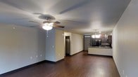 This single-floor residence offers a practical and comfortable layout designed for easy everyday living. The main living area is spacious and open, highlighted by rich, dark hardwood-style flooring that extends throughout the space, creating a warm and cohesive aesthetic. A ceiling fan with an integrated light fixture anchors the living room, providing both air circulation and ambient lighting. The open-concept design flows seamlessly into the kitchen, which features dark cabinetry, a functional peninsula with counter seating, and an elegant chandelier overhead that adds a refined touch. The kitchen is well-equipped with appliances including a refrigerator and stovetop, making it both stylish and practical. Along the back wall, multiple doorways lead to additional rooms, including what appears to be a closet and other private spaces, suggesting a thoughtful floor plan. The neutral wall tones throughout complement the darker flooring, lending the interior a balanced, understated look. Dark baseboards provide a clean contrast and a finished appearance to each room. Overall, this residence combines functional design with inviting details, making it a comfortable and well-appointed living space.