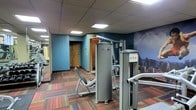 This fitness center offers a comprehensive workout environment designed to meet a range of exercise needs. The space is anchored by a selection of professional-grade strength-training machines, including a prominent multi-station cable and weight machine, providing users with options for full-body resistance training. A dedicated sauna room, accessible via a solid wood door, adds a wellness and recovery dimension to the facility.

The interior design features blue-painted walls accented by a striking cityscape mural, adding visual depth and an energizing atmosphere to the workout environment. The colorful, patterned carpet tiles covering the floor contribute a lively aesthetic while providing a durable and practical surface underfoot. Overhead, a drop ceiling with recessed flat panel lighting ensures the space is evenly and adequately illuminated throughout.

Wall-mounted mirrors extend the visual sense of the space and allow users to monitor their form during exercise. The single-floor layout is efficiently organized to keep equipment accessible without crowding, supporting a comfortable and functional workout experience. Overall, the fitness center strikes a balance between practicality and a motivating atmosphere suited for both casual and dedicated fitness routines.