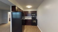 This single-floor residence offers a practical and comfortable layout designed for everyday living. The kitchen is a standout feature, outfitted with rich dark espresso cabinetry, light granite-style countertops, and a full suite of black appliances including a refrigerator, electric range, and over-the-range microwave. Durable wood-look flooring runs throughout the kitchen, providing an easy-to-maintain surface that complements the warm cabinetry tones. The living area is generously sized, finished with neutral-toned carpet and painted in soft gray tones. A front entry door and window allow natural light to fill the space, creating a bright and welcoming atmosphere. Two bedrooms are each carpeted and finished with the same neutral gray palette, offering a calm and restful environment. One bedroom includes a closet and connects to an adjoining room, while both benefit from ceiling-mounted light fixtures. The bathroom features stone-look tile flooring, a dark vanity with a granite-style countertop, white subway tile surrounding the tub, and stainless steel grab bars for added accessibility and safety. Throughout the residence, clean white trim and consistent finishes create a cohesive and move-in-ready aesthetic.