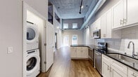 This two-floor urban loft seamlessly blends industrial character with modern finishes across a generously sized layout. The main level serves as the heart of the home, anchored by a well-appointed kitchen featuring white shaker cabinetry, dark granite countertops, stainless steel appliances, and a subway tile backsplash. A stacked in-unit washer and dryer are conveniently tucked nearby. Soaring exposed concrete ceilings with visible ductwork and track lighting reinforce the loft's industrial aesthetic throughout, while warm hardwood-style flooring provides a grounding contrast. The living area benefits from natural light streaming through large windows, with views of the surrounding brick building exterior. A private outdoor patio extends the living space, offering a tiled terrace framed by classic urban brick architecture. The upper floor houses the bedroom, a comfortably sized retreat with exposed concrete ceilings, industrial column accents, and dual windows that bring in natural light. A dedicated walk-in closet with full rod and shelf systems provides ample storage. The bathroom is finished with white cabinetry, a dark granite vanity top, large mirror, and a glass-enclosed shower, completing this refined urban living space.