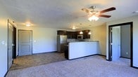 This single-floor space offers an open-concept layout that seamlessly connects the living area to the kitchen, creating a cohesive and functional environment. The living area is finished with plush wall-to-wall carpeting and is illuminated by a ceiling fan with integrated lighting, contributing to a comfortable and well-lit atmosphere.

The kitchen is designed for both style and practicality, featuring rich dark cabinetry that provides ample storage and a striking contrast against the light-toned walls. A built-in peninsula with a granite-style countertop serves as a breakfast bar, acting as a natural divider between the kitchen and living spaces. Stainless steel appliances, including a full-size refrigerator and microwave, lend a modern touch to the culinary space. Pendant lighting above the peninsula adds a warm, focused glow over the workspace.

The flooring transitions from tile in the kitchen and entry areas to carpet in the living space, clearly defining each zone within the open plan. The textured ceiling and neutral wall palette keep the space feeling bright and versatile. A doorway visible along the side wall suggests additional rooms beyond, completing the functional layout of this well-appointed single-floor residence.