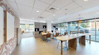 This thoughtfully designed single-floor commercial amenity space offers a generous, open layout across several distinct functional zones. The interior showcases a contemporary aesthetic, with a suspended grid ceiling outfitted with recessed lighting, large floor-to-ceiling windows, and neutral large-format tile flooring that transitions seamlessly throughout.

The main lounge area features a sophisticated arrangement of leather seating, swivel chairs, and a plush sofa grouping centered around a flat-screen display mounted on a wood-paneled accent wall. Adjacent to this is a lively recreation zone, complete with a foosball table and additional leather lounge chairs, all bathed in natural light from expansive glazed walls overlooking outdoor greenery.

A stylish café and dining area anchors one side of the space, featuring marble-top bistro tables, leather bar stools, a bold patterned accent wall, and a full-service kitchenette with stainless steel appliances, a clean white quartz countertop, and wood-grain cabinetry. An open bar-height counter island provides additional casual seating.

Warm wood shelving units with open display niches serve as functional dividers, adding texture and visual depth throughout. The abundant glazing ensures natural light permeates the entire space, creating an inviting and energetic atmosphere well-suited for collaboration, socializing, and daily amenity use.