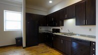 This single-floor space offers a thoughtfully designed interior that balances functionality with style. The kitchen is a standout feature, outfitted with rich, dark espresso-toned cabinetry that extends to the ceiling, maximizing storage capacity. Granite countertops in a speckled neutral tone provide durable and attractive work surfaces, while the full suite of black appliances — including a refrigerator, gas range, over-the-range microwave, and dishwasher — lends a cohesive, modern aesthetic. An under-cabinet light above the sink area adds practical task lighting, and a stainless steel double-basin sink completes the workspace. Throughout the space, warm-toned hardwood flooring adds character and a sense of continuity, its natural grain and patina giving the interior a timeless quality. The open layout adjacent to the kitchen provides a versatile area that benefits from a window allowing natural light to filter in, brightening the otherwise neutral white walls. Exposed piping along the upper wall adds an industrial design element that hints at the building's architectural character. Crown molding details further elevate the overall finish of the space, contributing to a polished yet approachable ambiance.