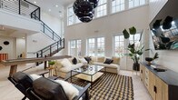 This expansive, open-concept space is defined by its dramatic double-height ceilings and an abundance of natural light streaming through oversized multi-pane windows. The main living area serves as the centerpiece, anchored by a shiplap feature wall with a mounted flat-screen TV, plush sectional seating, and a bold patterned area rug. Statement globe pendant lights in matte black hang from the soaring ceiling, adding a contemporary edge. A sleek metal-and-wood staircase with industrial railings leads to an upper mezzanine-level overlook, while structural white columns punctuate the open floor plan with architectural elegance. The bar area is thoughtfully designed with a dark cabinetry base, white quartz countertop, bar stool seating, and a cluster of gold globe pendant lights that cast a warm ambiance. Light herringbone-pattern hardwood flooring flows seamlessly throughout the entire space, unifying each zone. A dedicated games room features a full-size pool table alongside additional seating and entertainment options. The crisp white walls, tasteful greenery, and carefully curated mix of natural wood and black metal accents create a sophisticated yet welcoming atmosphere throughout this versatile space.