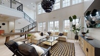 This expansive, open-concept space is defined by its dramatic double-height ceilings and an abundance of natural light streaming through oversized multi-pane windows. The main living area serves as the centerpiece, anchored by a shiplap feature wall with a mounted flat-screen TV, plush sectional seating, and a bold patterned area rug. Statement globe pendant lights in matte black hang from the soaring ceiling, adding a contemporary edge. A sleek metal-and-wood staircase with industrial railings leads to an upper mezzanine-level overlook, while structural white columns punctuate the open floor plan with architectural elegance. The bar area is thoughtfully designed with a dark cabinetry base, white quartz countertop, bar stool seating, and a cluster of gold globe pendant lights that cast a warm ambiance. Light herringbone-pattern hardwood flooring flows seamlessly throughout the entire space, unifying each zone. A dedicated games room features a full-size pool table alongside additional seating and entertainment options. The crisp white walls, tasteful greenery, and carefully curated mix of natural wood and black metal accents create a sophisticated yet welcoming atmosphere throughout this versatile space.