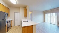 This single-floor residence offers a thoughtfully designed open-concept layout that seamlessly connects the kitchen and living areas. The kitchen is equipped with warm-toned wood cabinetry, a peninsula with a white countertop and undermount sink, and black appliances including a refrigerator, electric range, and microwave. Light wood-style flooring runs throughout the main living areas, contributing to a bright and clean aesthetic. The living area benefits from natural light through a large window with access to an outdoor balcony, along with a wall-mounted air conditioning unit for climate control. A dedicated in-unit laundry closet houses a full-size washer and dryer alongside wire shelving for added storage convenience. The bedroom features plush carpeting and ample wall outlet placement, providing a comfortable and functional space. The bathroom includes a wide vanity with a white countertop, an undermount sink with chrome fixtures, and a full bathtub and shower combination. Additional storage is available through multiple closets, including a pantry-style closet in the kitchen area. The overall color palette of soft beige and white tones throughout creates a clean, neutral, and welcoming atmosphere.