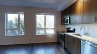This single-floor residence offers a clean, modern aesthetic highlighted by thoughtful design and quality finishes throughout. The kitchen is a standout feature, outfitted with sleek, dark wood cabinetry that extends to the ceiling for ample storage, complemented by light-toned countertops that create a striking contrast. Stainless steel appliances, including a built-in microwave, electric range, and oven, add a polished, contemporary touch. The rich, dark hardwood-style flooring runs throughout the space, grounding the interior with warmth and cohesion. Large windows flood the open area adjacent to the kitchen with natural light, offering pleasant views of mature trees outside. The neutral wall tones keep the space feeling fresh and versatile, suitable for a variety of furnishing styles. The open layout between the kitchen and the adjoining living area promotes a sense of spaciousness and easy flow. Clean lines, modern hardware, and well-placed lighting contribute to the overall contemporary ambiance, making this a functional and visually appealing single-level space.