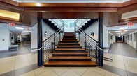 This two-floor space makes a striking first impression with its grand central staircase, a standout architectural feature finished in rich hardwood treads and framed by sleek black metal railings and bold cylindrical columns. A deep wood-paneled overhead beam crowns the entry area, adding warmth and sophistication to the tiled ground floor. The staircase ascends to an open upper level that overlooks the lower floor through the same refined metal railing system, creating a strong sense of vertical connectivity throughout the space.

The upper level opens into an inviting lounge area bathed in natural light from floor-to-ceiling windows framed in warm wood trim. These expansive windows offer views of the surrounding landscape and neighboring structures, giving the space an airy, well-connected feel. Seating areas feature a thoughtful mix of leather lounge chairs, teal upholstered furniture, and accent ottomans arranged to encourage relaxed gathering and conversation.

A kitchenette serves the space with white cabinetry, dark countertops, a stainless steel refrigerator, and a sink — accompanied by playful wall lettering that reads "But First, Coffee." The lower level corridors feature wood-look flooring, recessed lighting, and access to additional functional areas. Throughout both floors, the interplay of hardwood accents, neutral walls, and contemporary fixtures creates a polished yet welcoming environment.
