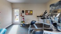 This fitness center is a compact yet fully outfitted exercise facility designed to support a well-rounded workout routine. The space features patterned carpeted flooring with teal accents and neutral-toned walls that create a clean, functional atmosphere. Natural light filters in through a window overlooking an outdoor pool and surrounding greenery, adding an energizing ambiance to the room.

The cardio equipment lineup includes treadmills and an upright stationary bike, each with integrated digital consoles for tracking workout metrics. An elliptical machine rounds out the cardiovascular offerings. For strength and functional training, the facility provides a rack of colorful kettlebells in varying weights, a wall-mounted medicine ball storage unit holding multiple weighted balls, resistance bands, TRX suspension straps, and foam yoga mats for stretching or floor exercises.

Bold graphic wall panels featuring athletic silhouettes and colorful geometric designs add a dynamic, motivating aesthetic to the space. A bulletin board near the entrance displays facility notices, and a fire extinguisher is conveniently mounted nearby for safety. The entry features a frosted-glass door, and an additional exterior door with views of the parking area provides easy access. A small refrigerator and wall-mounted equipment cabinet complete the amenities, making this fitness center a practical and engaging space for daily exercise.