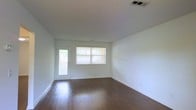 This single-story space presents a clean, minimalist living area designed with functionality and simplicity in mind. The room is finished with rich, dark hardwood flooring that contrasts sharply against the crisp white walls, creating a visually balanced and welcoming atmosphere. A textured popcorn ceiling adds subtle dimension overhead, while a centrally placed HVAC vent ensures comfortable climate control throughout the space.

Natural light is a defining feature of this room, sourced from a horizontal window fitted with white horizontal blinds, allowing for adjustable light control. Adjacent to the window, a white-framed exterior door with a glass panel further enhances the brightness of the space and provides a view of the surrounding greenery visible through the glass. White baseboards run cleanly along the perimeter of the room, adding a polished, finished look to the overall aesthetic.

The open layout offers a versatile, unobstructed floor plan well-suited for a variety of furniture arrangements. The neutral color palette and natural light give the space an airy, open quality, making it feel welcoming and adaptable to different interior design preferences.