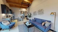 This single-floor unit showcases a striking loft-inspired design defined by soaring ceilings with exposed wooden beams and visible industrial elements, lending the space a warm yet contemporary character. The open-concept living area flows seamlessly from the entry, where large windows flood the room with natural light and offer views of the surrounding neighborhood. The living space is generously proportioned, accommodating a full seating arrangement alongside a dedicated reading nook with additional window exposure.

The kitchen is well-appointed with stainless steel appliances, including a range, microwave, dishwasher, and refrigerator, complemented by dark upper cabinetry, light lower cabinets, and a classic white subway tile backsplash. A mobile island with a butcher block top provides flexible counter and dining space.

The bedroom benefits from the same soaring wood-paneled ceiling with an exposed beam, a ceiling fan, a window with exterior views, and a spacious sliding-door closet. A second sitting area offers a quiet retreat with two tufted armchairs.

The bathroom is clean and modern, featuring a white vanity with integrated sink, gray tile shower-tub combination, and tasteful contemporary fixtures throughout.