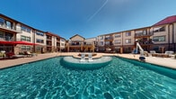 Located at 500 Apex Dr in Windsor, Colorado, this resort-style pool and spa area serves as a premier outdoor amenity space designed for relaxation and recreation. At the heart of the space is a generously sized swimming pool with crystal-clear blue water, featuring built-in lounge chairs positioned within the pool for a true resort experience. Adjacent to the pool, a raised circular spa provides a dedicated area for hydrotherapy and relaxation. The expansive concrete pool deck is outfitted with chaise lounges and large red market umbrellas, offering ample sun and shade options for residents. A covered pergola with string lighting anchors one side of the courtyard, providing a shaded retreat with additional seating. A separate fire pit lounge area features cushioned outdoor furniture arranged around a central fire table, ideal for cooler Colorado evenings. The entire pool area is enclosed by a secure black iron fence with stone-accented pillars and a gated entry. The surrounding multi-story building with balconies frames the courtyard, creating a private, enclosed atmosphere. The combination of warm sandstone tones, brick accents, and open Colorado skies gives this outdoor amenity space a welcoming and upscale character.