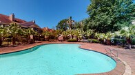 This amenity space features two distinct recreational areas designed to support both relaxation and active fitness. The outdoor pool area showcases a generously sized, freeform swimming pool with a classic turquoise finish, framed by a brick-toned paver deck. Mature palm trees, dense tropical greenery, and manicured shrubs create a lush, resort-inspired atmosphere, while a perimeter fence encloses the space for privacy. Brick residential buildings are visible beyond the landscaping, lending a warm, community-oriented character to the setting.

The fitness center is a bright, well-appointed space benefiting from abundant natural light through large floor-to-ceiling glass doors and windows that offer direct views of the pool area. The interior features light-toned carpet and hardwood-style flooring, recessed lighting, and full-length mirrored walls that enhance the sense of openness. Equipment includes multiple treadmills, elliptical machines, a cable multi-station, weight benches, a comprehensive dumbbell rack, barbells, and a medicine ball collection. Built-in wooden shelving provides organized storage for workout towels and accessories. A wall-mounted television adds convenience for cardio sessions. Together, these amenities create a complete and functional recreational environment.