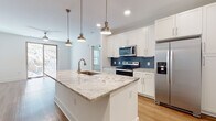 Located at 1040 Enclave Piney Lane in Asheville, NC, this well-appointed single-floor residence offers a clean, contemporary design throughout. The heart of the home is an open-concept kitchen featuring a large center island crowned with striking veined granite countertops and an undermount sink with a brushed gold faucet. Pendant lighting in a warm brass finish hangs above the island, adding an elegant touch. White shaker-style cabinetry lines the walls, complemented by a bold blue tile backsplash and a full suite of stainless steel appliances, including a side-by-side refrigerator, electric range, and over-the-range microwave. Warm-toned hardwood flooring flows throughout the main living and kitchen areas, creating a cohesive and inviting atmosphere. The bedrooms are finished with plush neutral carpeting and feature multiple windows that invite generous natural light and views of the surrounding landscape. The bathrooms are well-designed, with one offering a soaking tub and shower combination alongside wood-look flooring and direct access to a walk-in closet. A second bathroom features a glass-enclosed walk-in shower and granite vanity countertop. Throughout the residence, bright white walls, clean lines, and recessed lighting contribute to a fresh, modern ambiance.