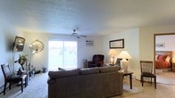 This single-floor residence offers a welcoming and functional layout centered around a spacious living area. The living room is comfortably furnished with a large sofa and a reclining armchair, providing ample seating for relaxation. Soft carpet flooring runs throughout, adding warmth to the space. A sliding glass door fills the room with natural light and offers a view of the outdoors, creating an open and airy atmosphere. A ceiling fan is centrally positioned above, ensuring year-round comfort alongside a wall-mounted air conditioning unit. Warm table lamps placed around the room contribute to a cozy ambiance during evening hours. The neutral-toned walls provide a clean, understated backdrop that complements the furnishings. A decorative wall clock and framed wall art add personal character to the space. The overall design is practical and inviting, with a straightforward layout that maximizes the use of the available floor plan. The connection between the interior living space and the outdoor area via the sliding glass door is a standout feature, enhancing both the sense of space and the flow of natural light throughout.