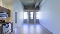 This single-floor space makes a striking first impression with its bold juxtaposition of industrial and contemporary design elements. The most notable architectural feature is the exposed concrete ceiling, complete with visible structural beams, which lends the space a raw, loft-like character. Elevated ceiling height adds a sense of volume and openness throughout the interior.

The main living area is bright and airy, bathed in natural light streaming through a set of arched windows framed by sheer white curtains. These distinctive arched window frames serve as a charming architectural detail, drawing the eye outward while connecting the interior to the surrounding urban environment. The light hardwood flooring runs seamlessly throughout, adding warmth that beautifully contrasts the cooler tones of the concrete above.

The walls are finished in a clean, neutral palette, keeping the space feeling open and versatile. A doorway visible from the main area hints at additional rooms or spaces beyond, contributing to a functional and well-connected floor plan. Overall, the space strikes a thoughtful balance between rugged industrial character and polished modern living, making it a compelling and unique environment suited for a variety of uses.
