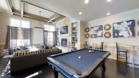 This single-floor entertainment space is thoughtfully designed to blend relaxed comfort with upscale amenities. At the forefront is a full-sized billiards table with a slate-blue felt surface, serving as the centerpiece of the room and setting a recreational tone. Behind it, a striking stone fireplace framed in stacked natural limestone anchors the wall, with a flat-screen television mounted above and flanked by white built-in shelving units displaying curated décor pieces and accessories.

Adjacent to the fireplace, a lounge area features plush seating with bold striped and patterned throw pillows in a neutral, grey, and yellow palette. A tray ceiling detail overhead adds architectural depth, while a modern chandelier provides ambient lighting. Recessed lighting throughout ensures the space is well-illuminated.

A dining or café-style seating area sits to the right, complemented by decorative sunburst mirrors and contemporary artwork on the walls. Dark hardwood flooring runs throughout, providing contrast against the light-toned walls and stone elements. Large windows along the rear wall invite natural light, enhancing the open and inviting ambiance of this versatile entertainment lounge.