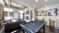This single-floor entertainment space is thoughtfully designed to blend relaxed comfort with upscale amenities. At the forefront is a full-sized billiards table with a slate-blue felt surface, serving as the centerpiece of the room and setting a recreational tone. Behind it, a striking stone fireplace framed in stacked natural limestone anchors the wall, with a flat-screen television mounted above and flanked by white built-in shelving units displaying curated décor pieces and accessories.

Adjacent to the fireplace, a lounge area features plush seating with bold striped and patterned throw pillows in a neutral, grey, and yellow palette. A tray ceiling detail overhead adds architectural depth, while a modern chandelier provides ambient lighting. Recessed lighting throughout ensures the space is well-illuminated.

A dining or café-style seating area sits to the right, complemented by decorative sunburst mirrors and contemporary artwork on the walls. Dark hardwood flooring runs throughout, providing contrast against the light-toned walls and stone elements. Large windows along the rear wall invite natural light, enhancing the open and inviting ambiance of this versatile entertainment lounge.