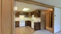 This single-floor space centers around a well-organized kitchen that features an L-shaped layout designed to maximize functionality. The kitchen is outfitted with dark brown wood cabinetry, both upper and lower, providing generous storage throughout. White appliances, including an electric range and dishwasher, complement the light-toned countertops, creating a clean contrast against the darker cabinet finish. A stainless-style sink is positioned along the countertop run, conveniently located beneath upper cabinets. The kitchen benefits from flush-mounted ceiling light panels that provide consistent, even illumination across the workspace. A drop ceiling with a textured tile finish runs throughout, giving the space a structured, uniform overhead appearance. The flooring transitions from a warm wood-look vinyl in the kitchen area, adding a touch of warmth to the overall palette. A structural beam detail is visible at the kitchen's entry point, framing the space distinctly. The overall ambiance is practical and straightforward, with a neutral color scheme that offers a solid foundation for personalization or renovation.