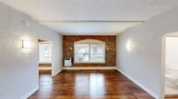 This single-floor unit offers a well-organized layout encompassing two living areas, a kitchen, a bathroom, a closet, and a hallway. The space is defined by its rich, dark-stained hardwood floors that flow continuously throughout, lending warmth and character to every room. A standout architectural feature is the exposed brick accent wall in the main living area, framing an arched window surround that adds a distinctive vintage charm. Natural light enters through multiple windows fitted with horizontal blinds, brightening the light gray walls and creating an inviting ambiance. Structural beams are visible along the ceiling, further enhancing the space's classic character. The two living areas are connected through an arched entryway, providing an open yet defined sense of flow. Wall sconces add a soft, layered lighting element throughout. The kitchen is outfitted with crisp white shaker-style cabinetry, a stainless steel gas range, and a undermount sink, all set against a clean white countertop and continuing hardwood flooring. The bathroom features floor-to-ceiling white subway tile surrounding a tub-shower combination and includes a window for natural ventilation and light. A dedicated closet with a wide opening provides practical storage. Overall, the space blends vintage architectural details with clean, updated finishes.