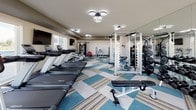 This fitness center offers a comprehensive workout environment designed to accommodate a wide range of exercise needs. The space is laid out across a single floor and is finished with distinctive multi-tone carpet tiles in shades of teal, beige, and gray, lending the room a modern, polished aesthetic.

The cardio section is well-appointed with multiple treadmills and stationary cycling bikes, each positioned to take advantage of the large windows that line the walls. These expansive windows flood the space with natural light and frame views of the surrounding cityscape, creating an energizing atmosphere during workouts. Wall-mounted flat-screen televisions are conveniently positioned throughout for entertainment.

The strength training area is equally well-equipped, featuring a multi-station cable machine, a power rack, a full dumbbell rack with a wide range of weights, and adjustable benches. A medicine ball rack and a BOSU balance trainer round out the functional fitness offerings. Full-length mirrors line one wall, enhancing the sense of openness and allowing users to monitor their form.

Modern circular ring-style light fixtures are mounted to the ceiling throughout, providing bright, even illumination across the entire space. The overall ambiance is clean, contemporary, and purpose-built for an effective fitness experience.