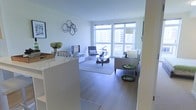 This thoughtfully designed single-floor residence offers a clean, contemporary aesthetic defined by an open-concept layout and an abundance of natural light. The living area serves as the heart of the space, featuring warm hardwood flooring that extends throughout, crisp white walls, and expansive floor-to-ceiling windows that frame sweeping urban views and flood the interior with daylight.

The living room is furnished with a comfortable, neutral-toned seating arrangement centered around a sculptural glass coffee table, complemented by a coordinating accent chair and a sleek arc floor lamp. A low-profile media console anchors one wall, while a striking geometric metal wall sculpture adds an artistic focal point to the room. A small side table in a bold green finish introduces a subtle pop of color to the refined palette.

The open layout flows seamlessly from the living space toward the kitchen area, separated by a bright white breakfast bar that provides casual seating and defines each zone without sacrificing the airy, connected feel of the home. The overall ambiance is light, modern, and welcoming — ideal for both everyday living and entertaining, with the urban cityscape serving as a dynamic backdrop.