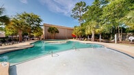 Located at 206 Cedar Branch Lane in Wilmington, North Carolina, this outdoor amenity space offers a well-appointed resort-style recreational area designed for relaxation and leisure. The centerpiece of the space is a generously sized freeform swimming pool with crystal-clear turquoise water, complemented by a connected shallow wading area — ideal for lounging or cooling off. The pool deck is finished in a smooth concrete surface, providing ample space for poolside furniture, including lounge chairs and vibrant green seating options positioned throughout the area.

A standout feature of this outdoor space is the hammock station, nestled beneath a canopy of mature trees, offering a shaded retreat for relaxation. A stone-accented outdoor grill area is also present, making the space suitable for outdoor gatherings. The entire area is enclosed by a sturdy black iron fence, ensuring security and privacy.

The landscaping is lush and tropical in character, with tall palm trees, mature pines, and dense greenery creating a serene, park-like atmosphere. Adjacent to the pool is a charming clubhouse-style building featuring a covered porch with columns and large windows, providing a welcoming gathering space. The combination of thoughtful amenities, natural surroundings, and Southern coastal charm makes this outdoor space a true highlight of the property.